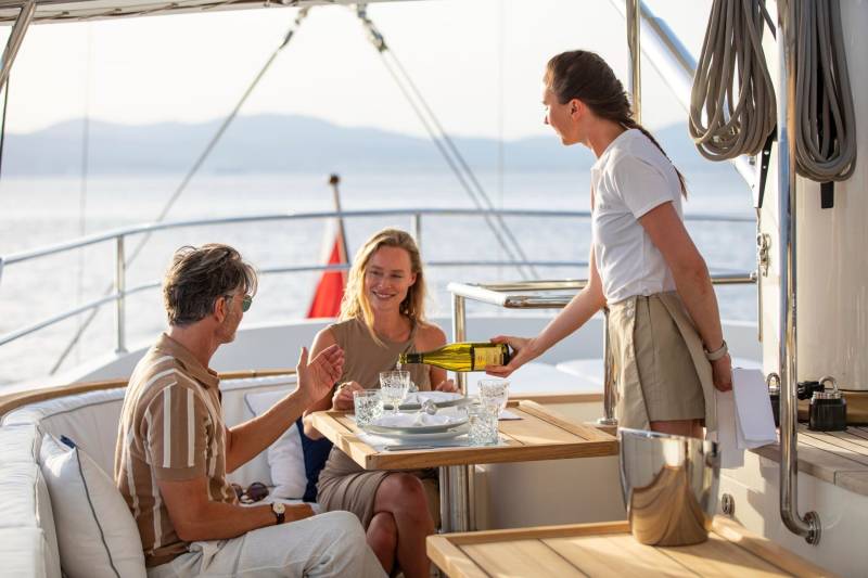 A couple enjoys a leisurely meal with wine being poured by a server on a luxurious yacht at sea.