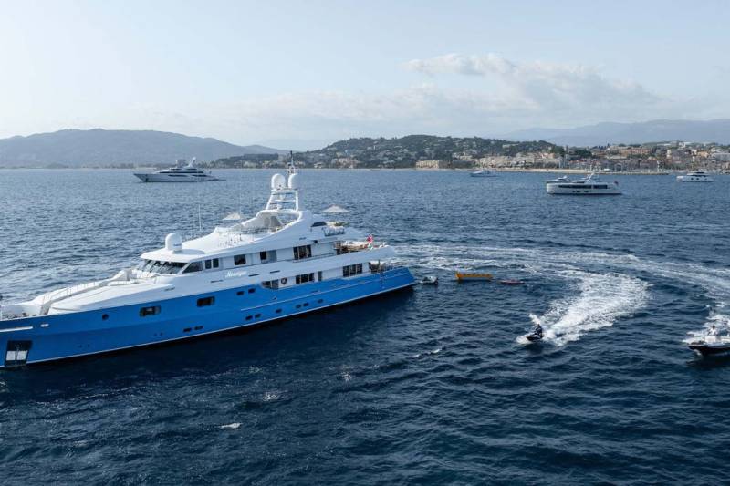 A vibrant blue and white superyacht anchors in the bay with jet skis and a tender nearby.