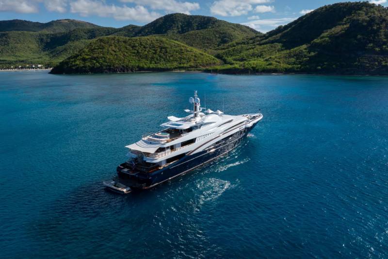 A luxurious superyacht cruises through the clear blue waters off a lush, green island.
