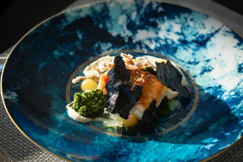 A beautifully plated seafood dish with vibrant green garnishes and dark crispy elements sits on a striking blue marbled plate.