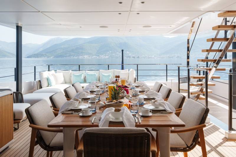 A beautifully set dining table awaits guests on a luxurious yacht deck, overlooking a serene bay with verdant mountains.