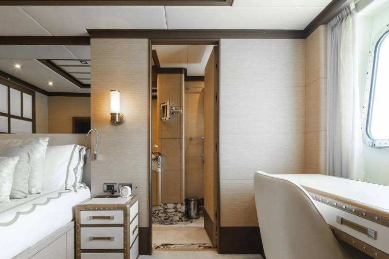 A luxurious yacht cabin features a comfortable bed, a sleek desk, and a glimpse into an elegant en-suite bathroom.
