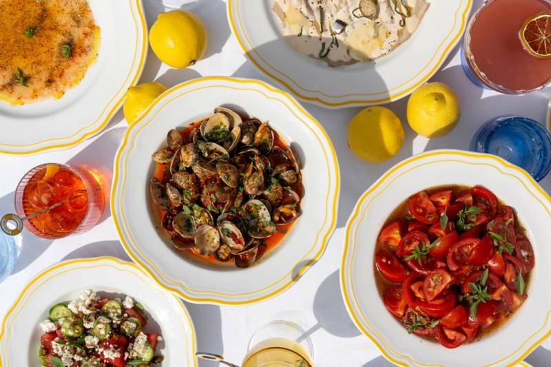 A vibrant spread of Italian dishes, including clams, carpaccio, and tomato salad, is set against a bright white tablecloth with lemons and drinks.