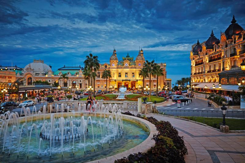 The opulent Casino de Monte-Carlo glows under a twilight sky, its grandeur reflected in the surrounding fountains and palm trees.
