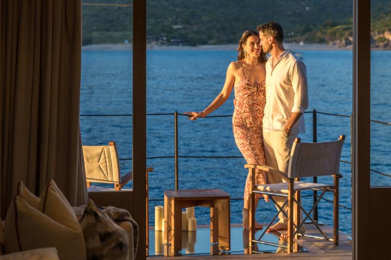 A couple embraces on a balcony overlooking a tranquil blue sea with a lush green hillside in the distance.