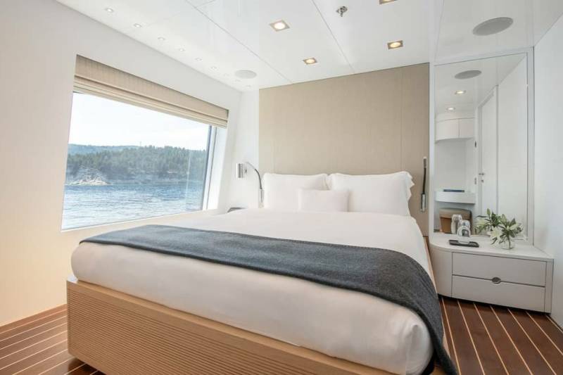 A modern bedroom with a large window offers stunning views of a tranquil body of water and lush green hills.