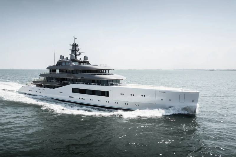 A sleek, modern superyacht cruises through calm waters under a bright, hazy sky.