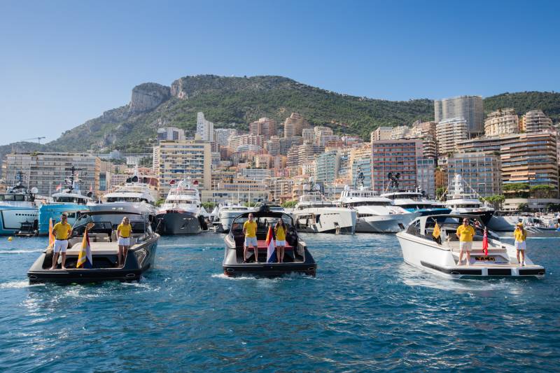 Luxury yachts cruise the azure waters of a Mediterranean harbor, with a densely populated hillside city as a backdrop.