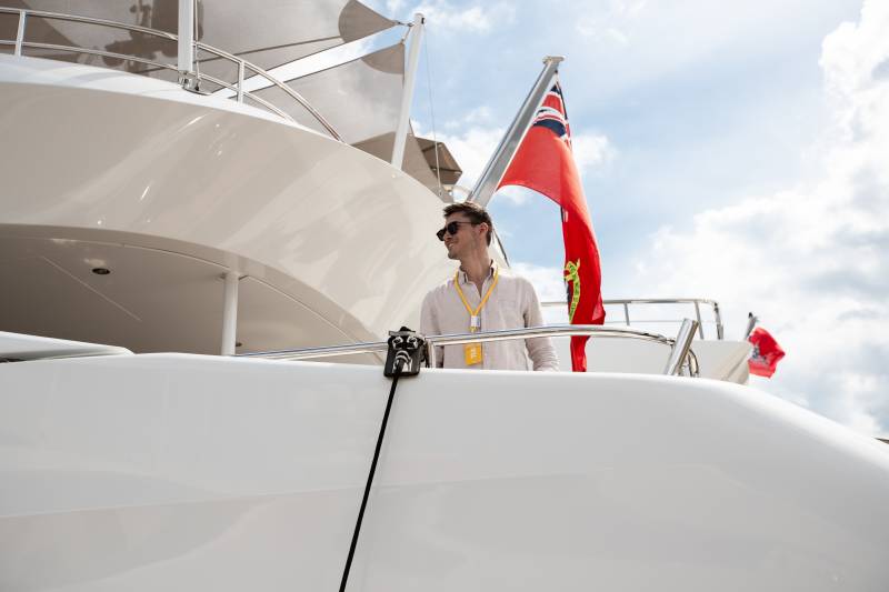 A person in a white shirt stands on the deck of a large white yacht, with a British flag waving in the background.