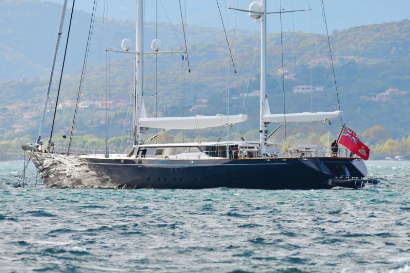 A majestic dark blue yacht with white sails glides across choppy turquoise waters under a hazy sky.