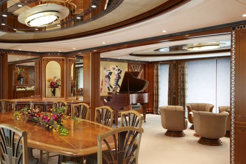 An opulent yacht dining room features a grand piano, elegant dining table, and plush seating, all adorned with rich wood and decorative art.
