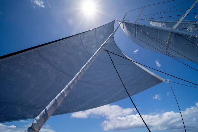 Sunlight streams through the billowing sails of a sailboat against a vibrant blue sky dotted with clouds.