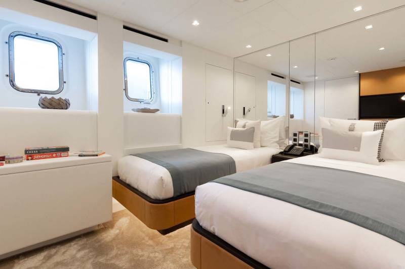 Twin beds with grey throws are neatly made in this bright, modern cabin featuring porthole windows and mirrored walls.