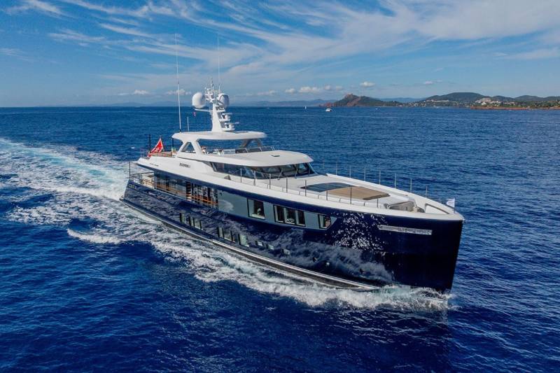 A sleek navy yacht glides through the azure waters under a bright blue sky with scattered clouds.