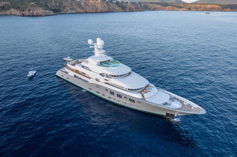 A luxurious superyacht glides through the deep blue Mediterranean Sea, with a rugged coastline in the background.