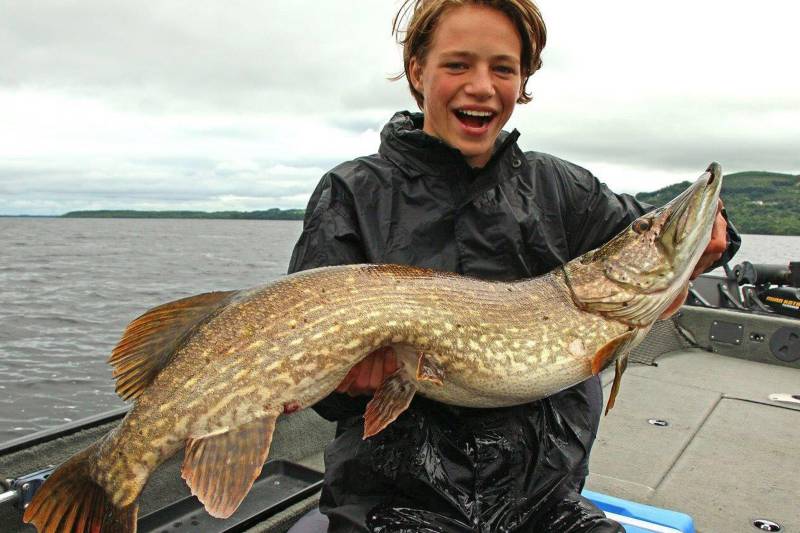 A beaming young angler proudly displays a massive pike caught from a boat on a cloudy day.