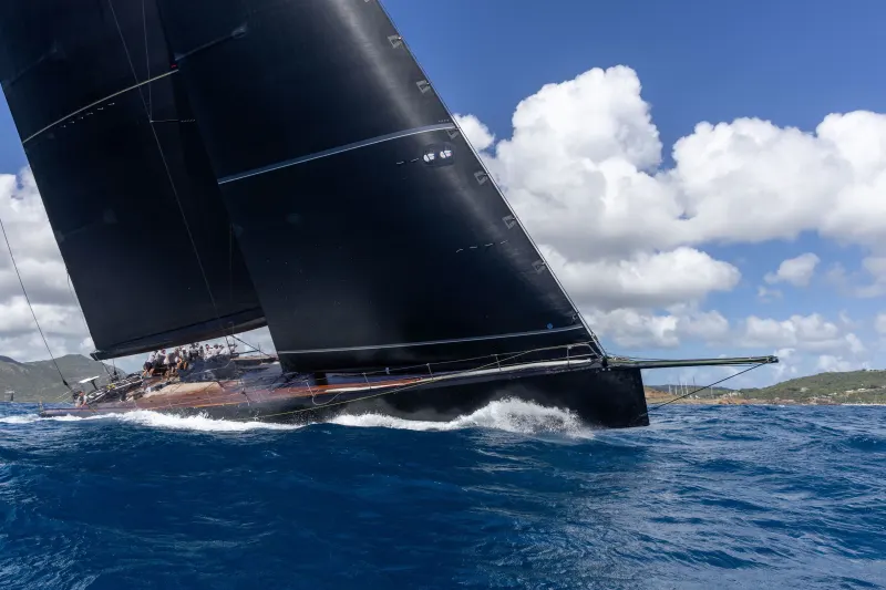 A sleek black sailboat cuts through the deep blue ocean, its sails billowing under a bright, cloudy sky.
