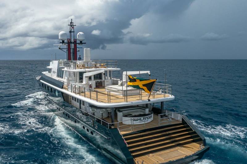 A luxurious yacht with a Jamaican flag sails through choppy blue waters under a dramatic cloudy sky.
