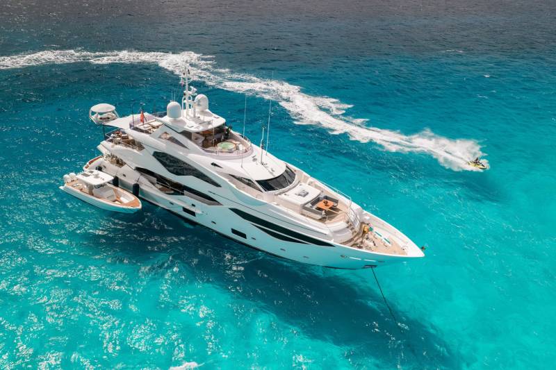 A large, luxurious yacht cruising through the crystal-clear, turquoise waters of what appears to be a tropical or subtropical location, with a wake of white foam trailing behind it.