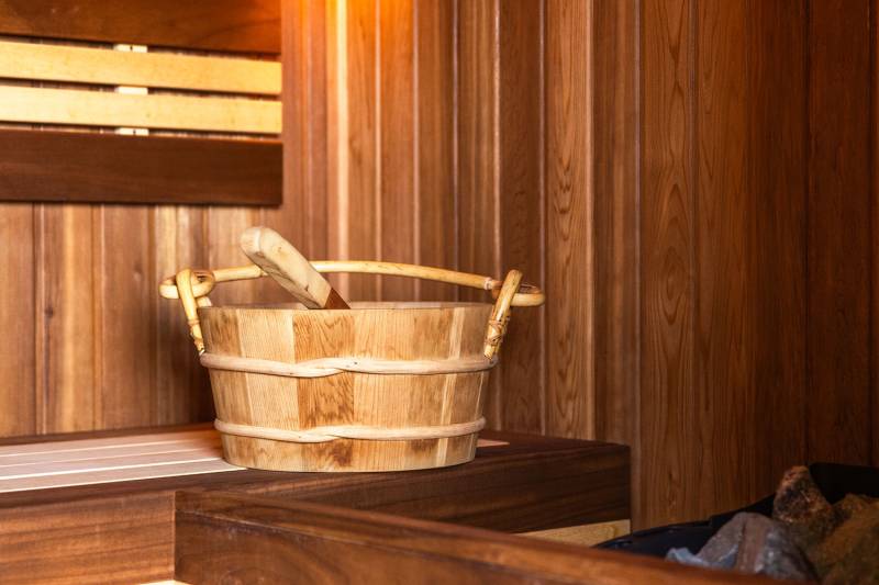 A wooden bucket and ladle rest on a bench in a warm, wood-paneled sauna.