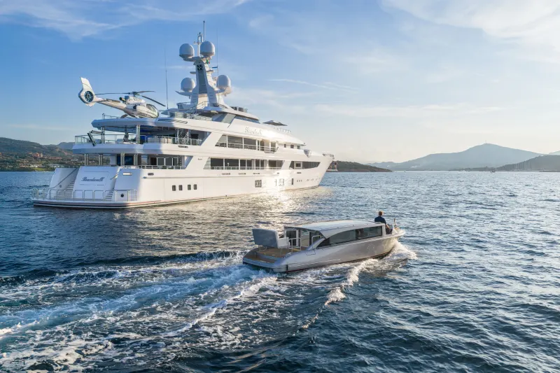 A sleek superyacht with a helicopter on deck cruises the deep blue waters near a verdant coastline.