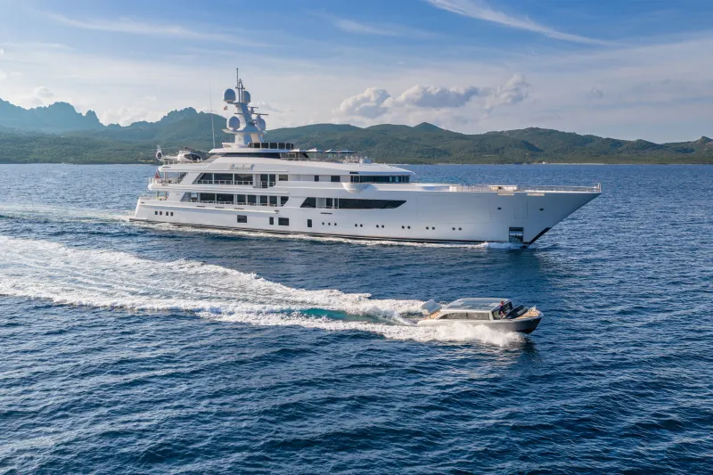 A luxurious superyacht cruises through the azure waters, leaving a frothy wake behind it, with a smaller tender boat in tow.