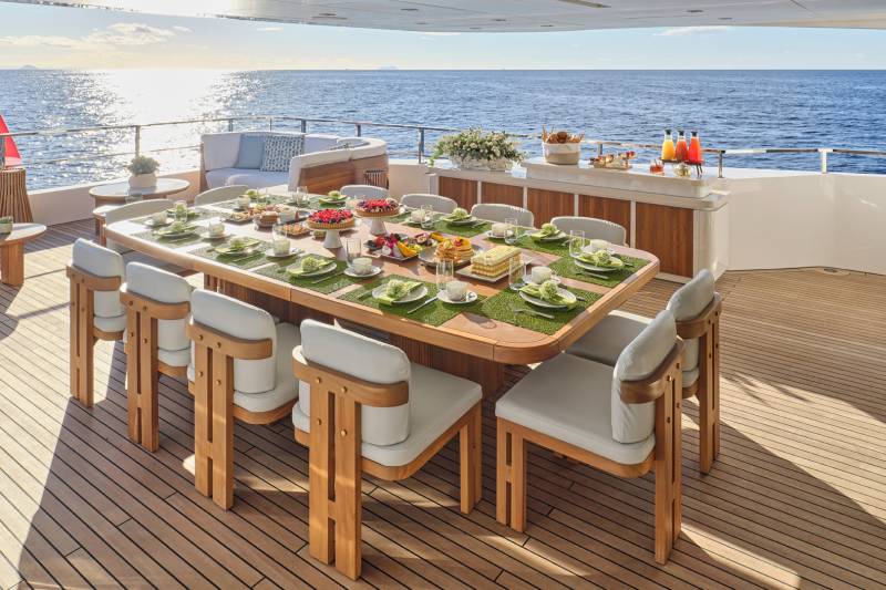 A luxurious outdoor dining area on the deck of a yacht, with a long wooden table laden with an array of appetizing dishes and surrounded by comfortable seating, all set against the backdrop of a serene ocean view.