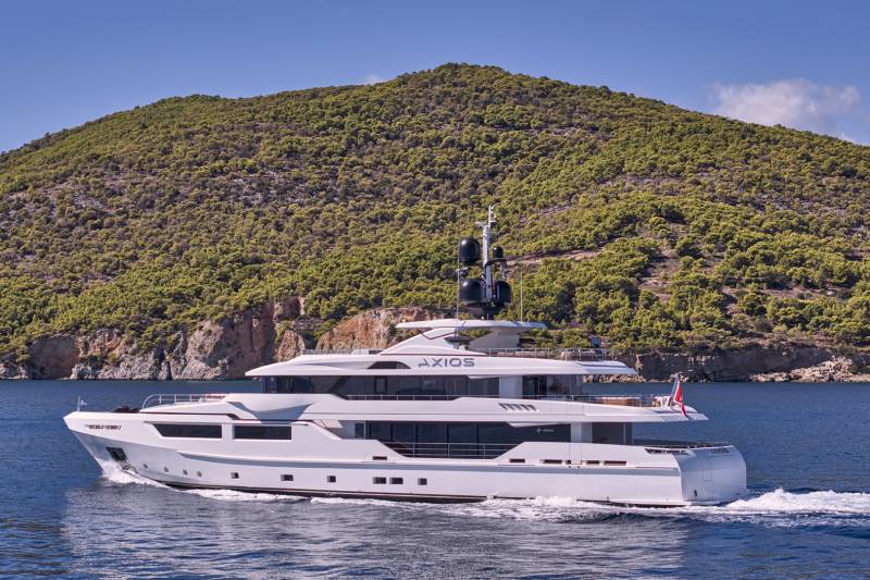A large, luxurious white yacht cruises through the calm, blue waters, with a mountainous, lush green landscape rising in the background.