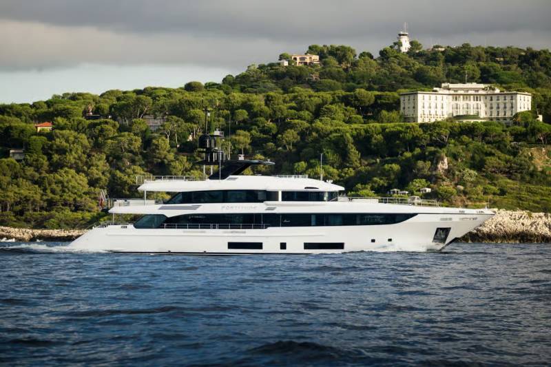 A sleek white yacht glides through the deep blue sea, with lush green hills and elegant buildings rising behind it.
