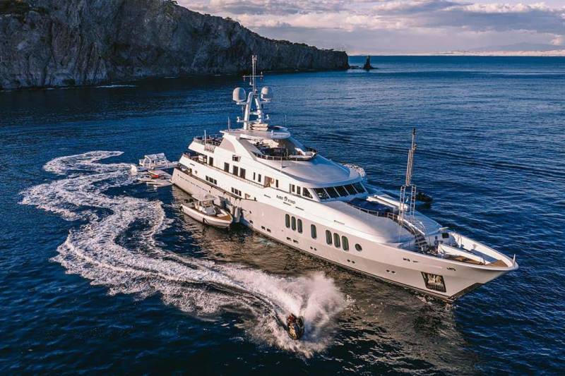 A luxurious superyacht cruises through the azure ocean, leaving a trail of white wake as a jet ski zips by.
