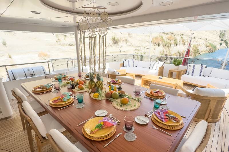 A beautifully set dining table on a yacht deck awaits a meal with a scenic backdrop of cliffs and water.