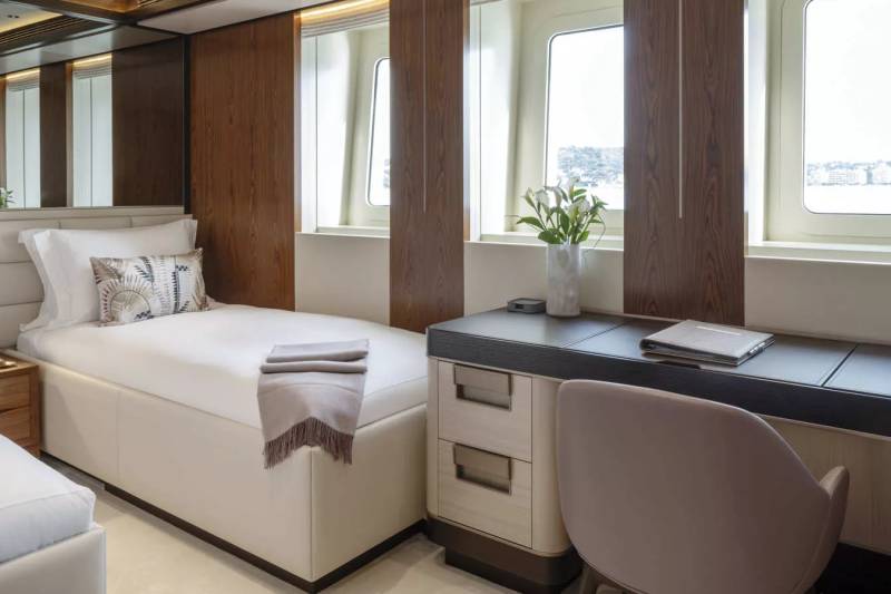 A modern yacht cabin features two twin beds, a sleek desk with a chair, and large windows overlooking a coastal landscape.