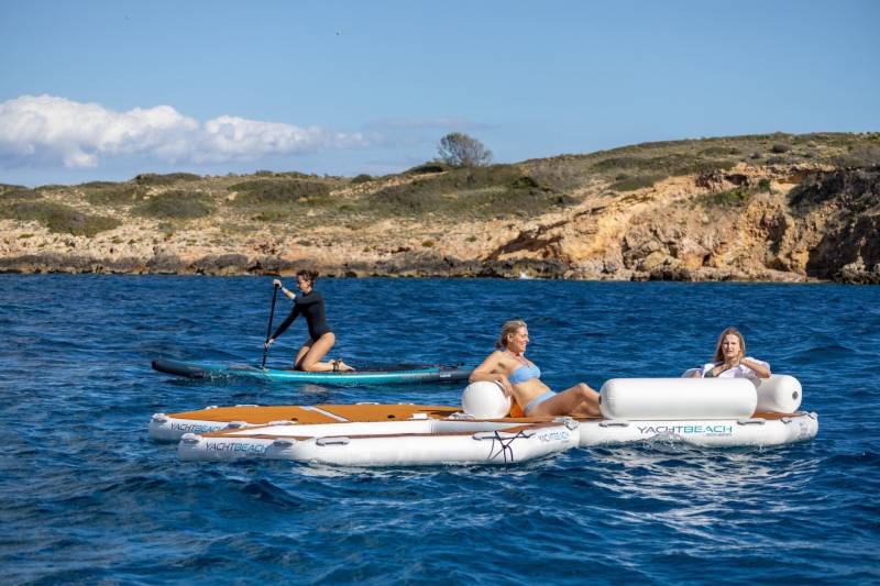 Two women relax on a floating platform while another paddles a stand-up paddleboard near a rocky coastline.