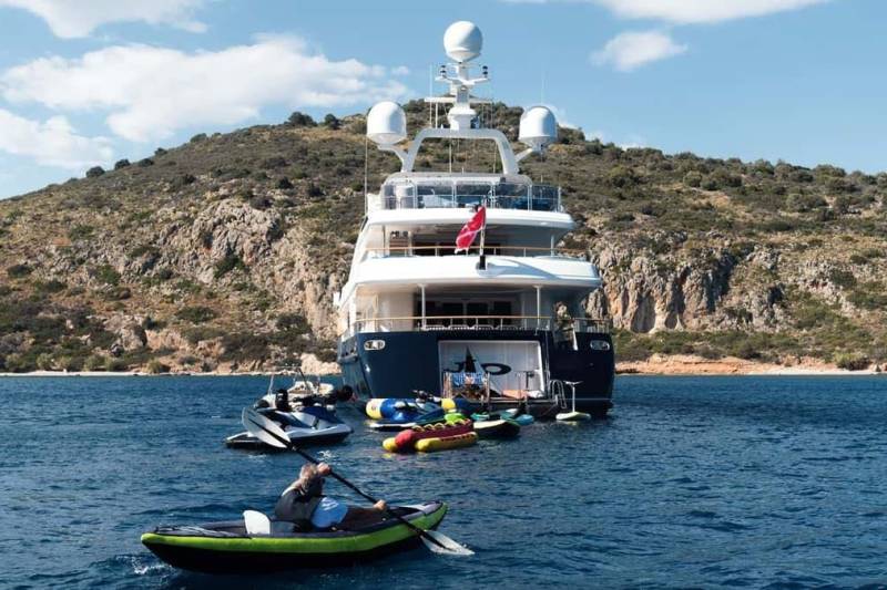 A person kayaks in the foreground with a large yacht and various water toys anchored near a rocky, tree-covered island.