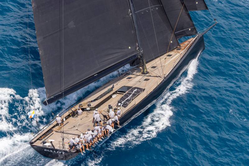 A sleek black sailboat cuts through the blue ocean waves with a crew of sailors on deck.
