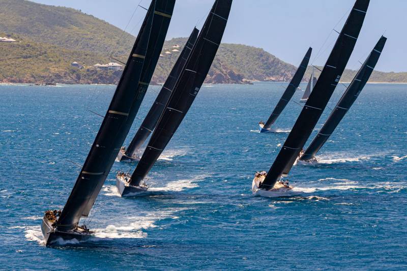 Sleek black sails dominate the azure waters as racing yachts vie for position under a clear blue sky.