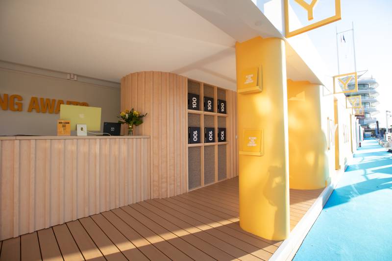 A bright yellow structure with wooden accents and a blue carpet creates a vibrant outdoor exhibition space.