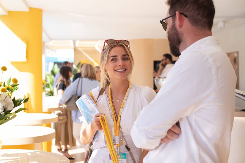 A woman with blonde hair and red sunglasses smiles while holding brochures and talking to a man with a beard.
