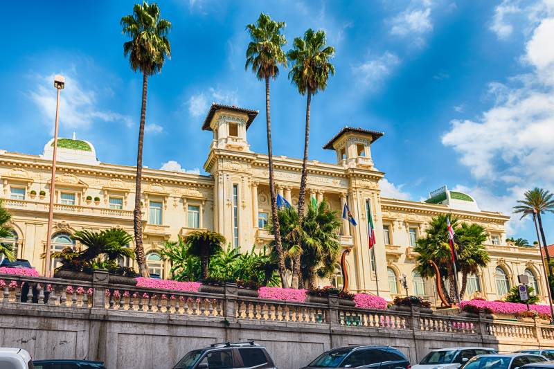 Palm trees and vibrant pink flowers frame the ornate facade of the historic Casino Municipale under a bright blue sky.