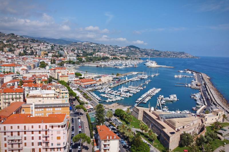 A picturesque Italian coastal town unfolds with a vibrant harbor filled with sailboats and a historic fortress overlooking the azure sea.