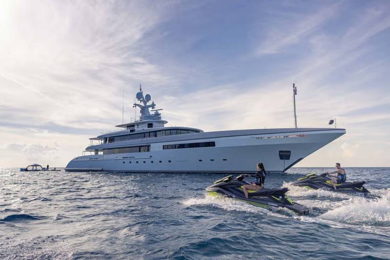 Two jet skiers enjoy a sunny day on the water with a large yacht in the background.