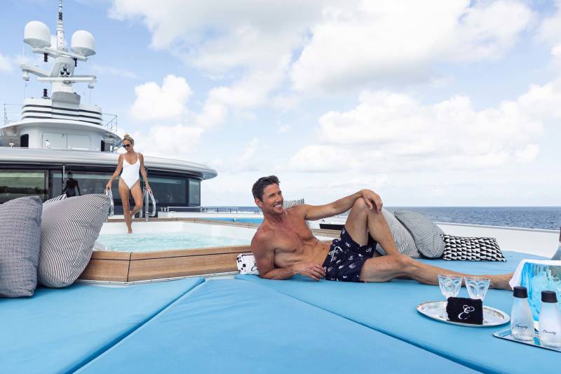 A man relaxes on a yacht's sundeck while a woman emerges from a hot tub on a bright, cloudy day.