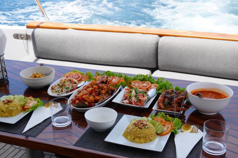 A delicious spread of Thai food is served on a yacht deck with the ocean in the background.