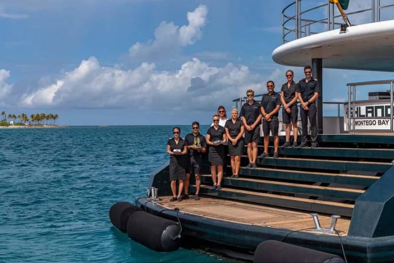 A yacht crew poses on the steps of their vessel with a tropical island in the background.