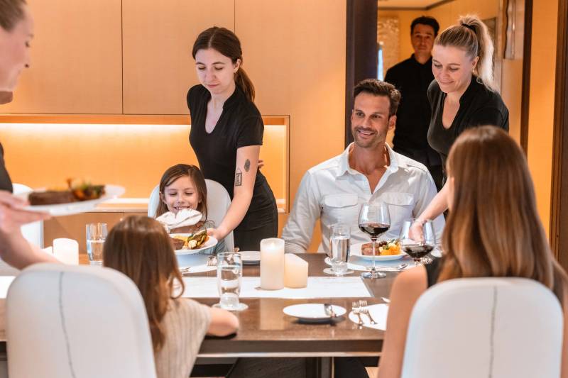 A family enjoys a formal dinner with attentive servers in a luxurious dining room.