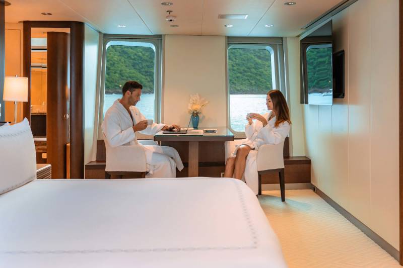 A couple enjoys coffee and a laptop in plush robes while overlooking a lush green landscape from their luxurious cabin.
