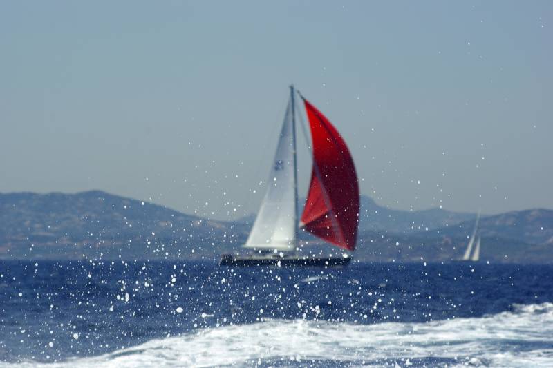 A sailboat with a red and white sail glides across the sparkling blue waters, surrounded by mountains in the distance.