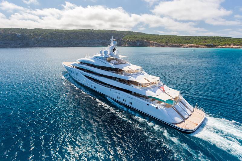 A luxurious white superyacht glides through the sparkling blue ocean near a verdant, hilly coastline.