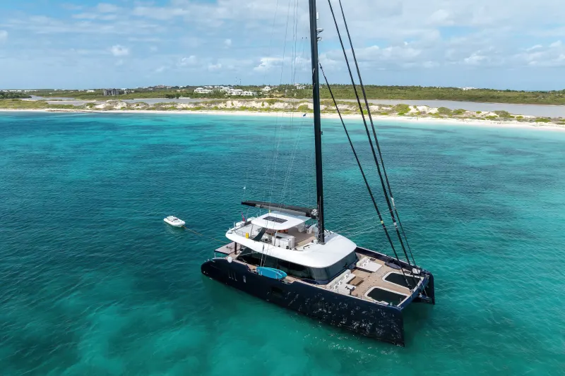 A large white catamaran sailboat is anchored in the crystal-clear turquoise waters of a tropical beach, with a lush green coastline visible in the background.