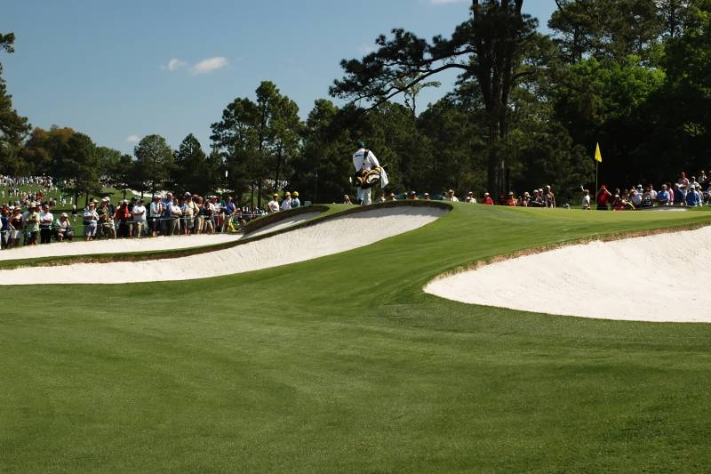 A golfer in a white uniform and green hat walks past a sand trap with spectators watching from the sidelines.
